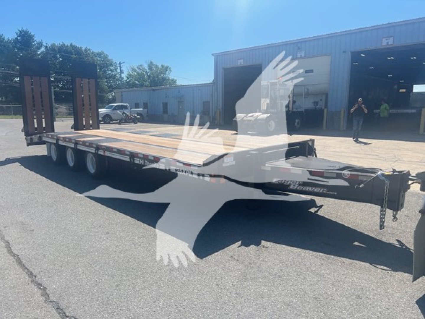 Trailer showing a long flatbed with dual rear axles and wooden deck; front tongue area with chains; Eager Beaver logo and model number not legible; open building in background.