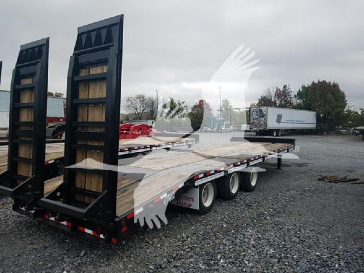 Trailer flatbed with ramps and dual rear tires, light-colored wood deck, in a gravel lot with other trailers in background.
