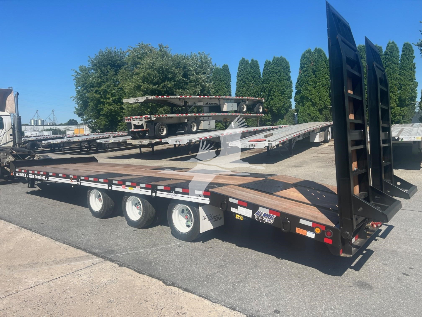 Trailer showing a lowboy deck and fold-up ramps with three axles in the foreground and another flatbed trailer in the background.