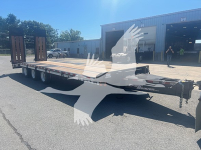 Trailer showing a long flatbed with dual rear axles and wooden deck; front tongue area with chains; Eager Beaver logo and model number not legible; open building in background.