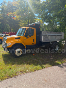 Dump truck showing orange International 7300 cab and grey dump body in a grassy lot with trees.