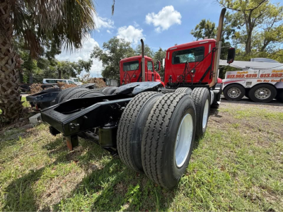Rear view of a red Mack CXU613 day cab truck chassis with dual rear axles and large tires. Text on distant trucks and a sign visible in background.