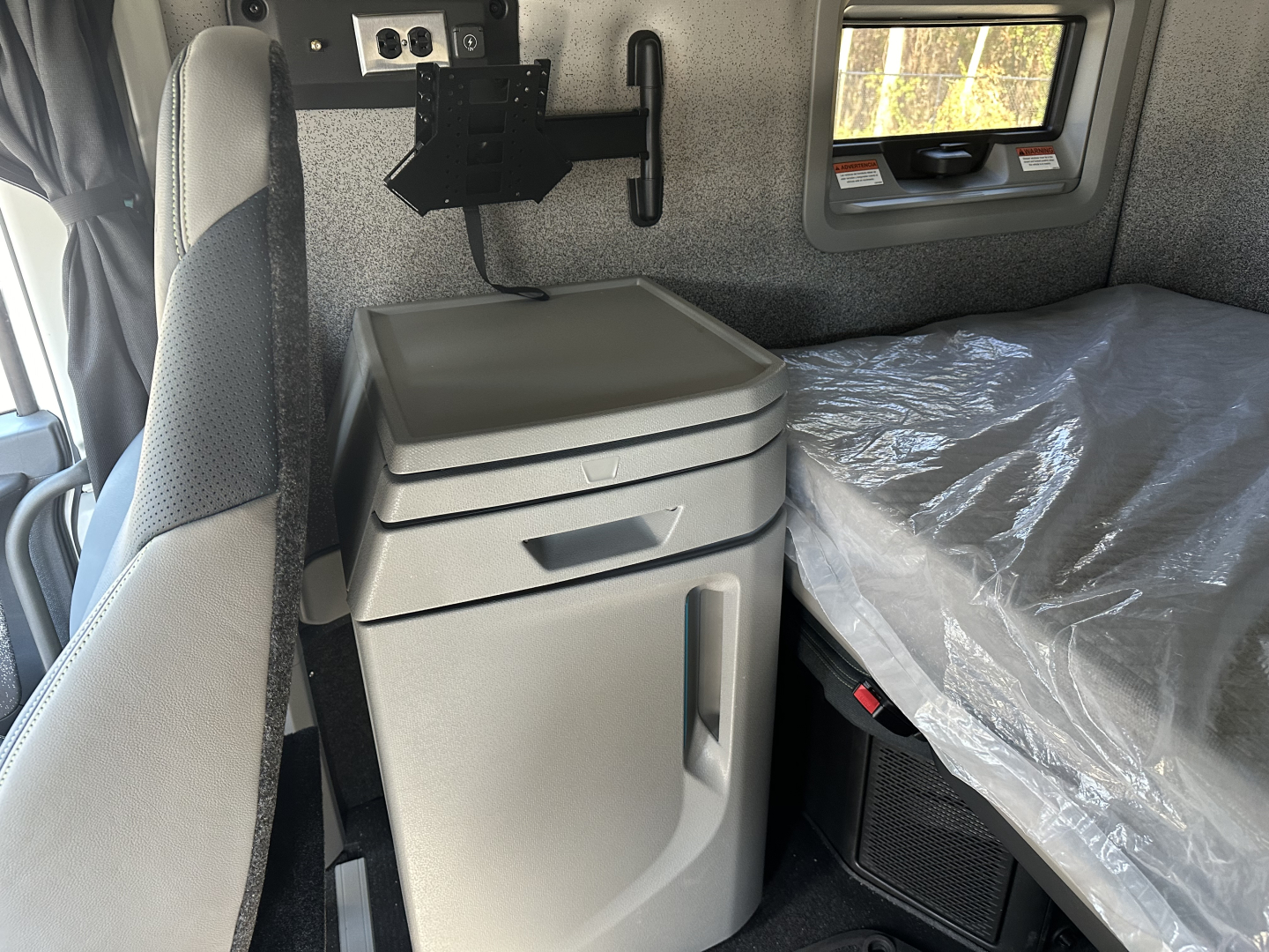 Volvo VNL64T860 sleeper cab interior showing bedside storage with grey fridge or cabinet, window, and bed with plastic cover; cab area with mounting bracket and panel.
