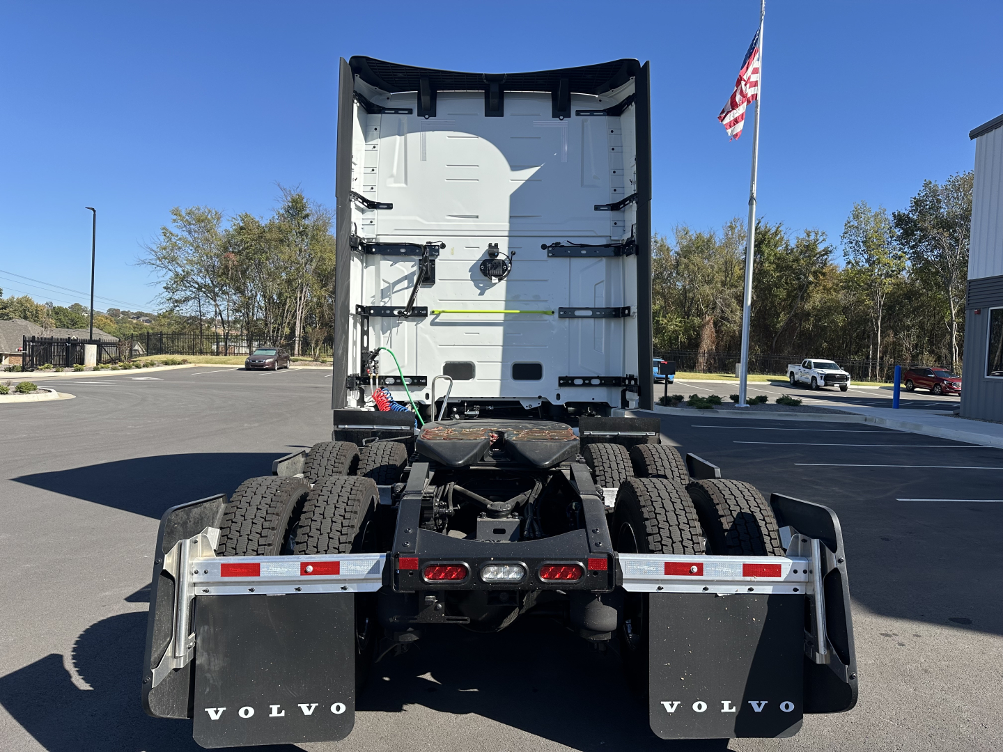 Truck cab rear view showing Volvo sleeper tractor frame and dual rear axles with parking lot and flagpole in background.