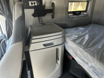 Volvo VNL64T860 sleeper cab interior showing bedside storage with grey fridge or cabinet, window, and bed with plastic cover; cab area with mounting bracket and panel.