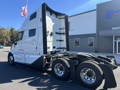 Volvo VNL64T860 tractor cab rear-left side showing sleeper, exposed fifth wheel, dual rear axles, chrome wheels; white exterior, dealership building in background.