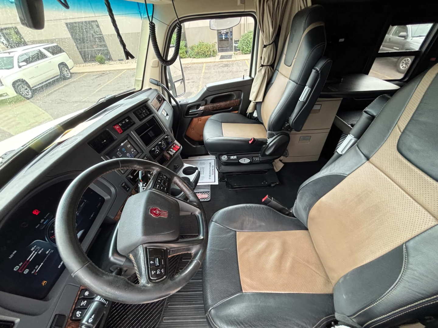 Truck cab interior of a Kenworth T680: steering wheel, dashboard, driver seat, passenger seat, center console, and door panel; visible upholstery in tan and black, multiple control panels, and gear lever.