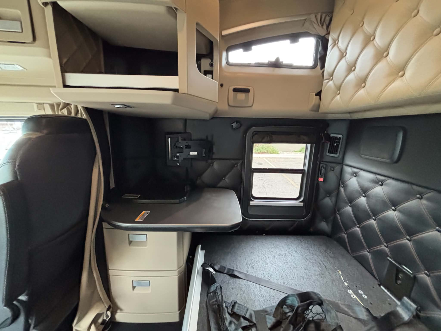 Truck cab interior of a 2024 Kenworth T680 showing sleeper area with quilted headboard, overhead storage, side window, and a fold-out desk/table.