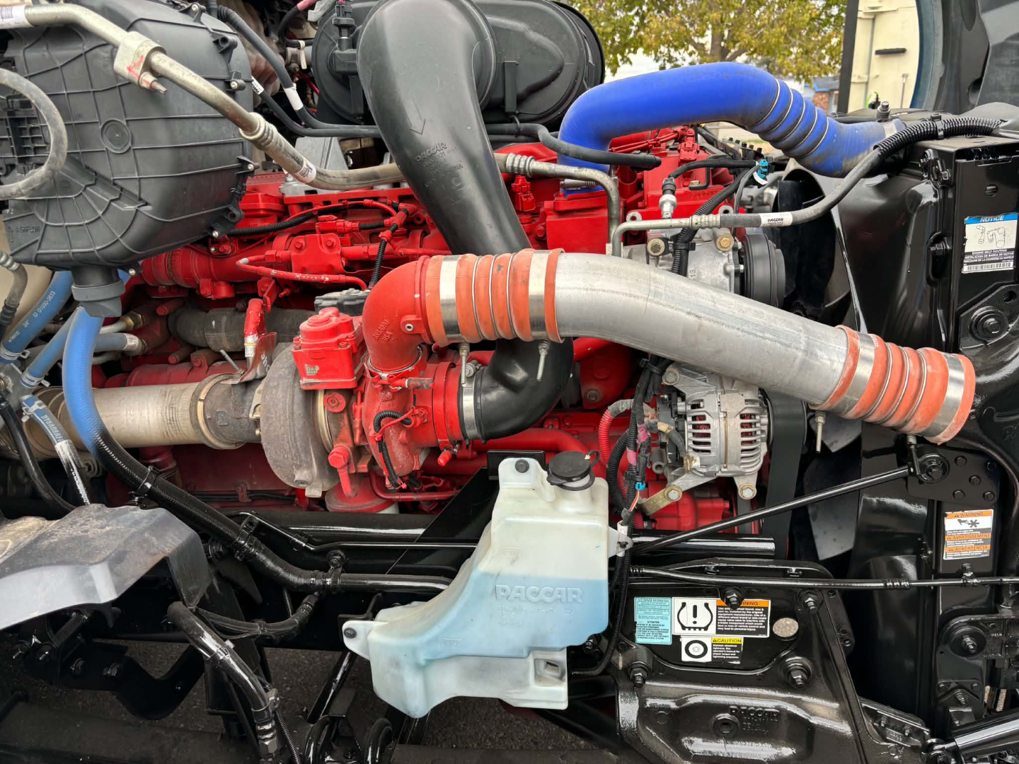 Kenworth T680 engine bay showing red engine block, blue coolant hose, gray radiator hoses, alternator, and coolant reservoir.