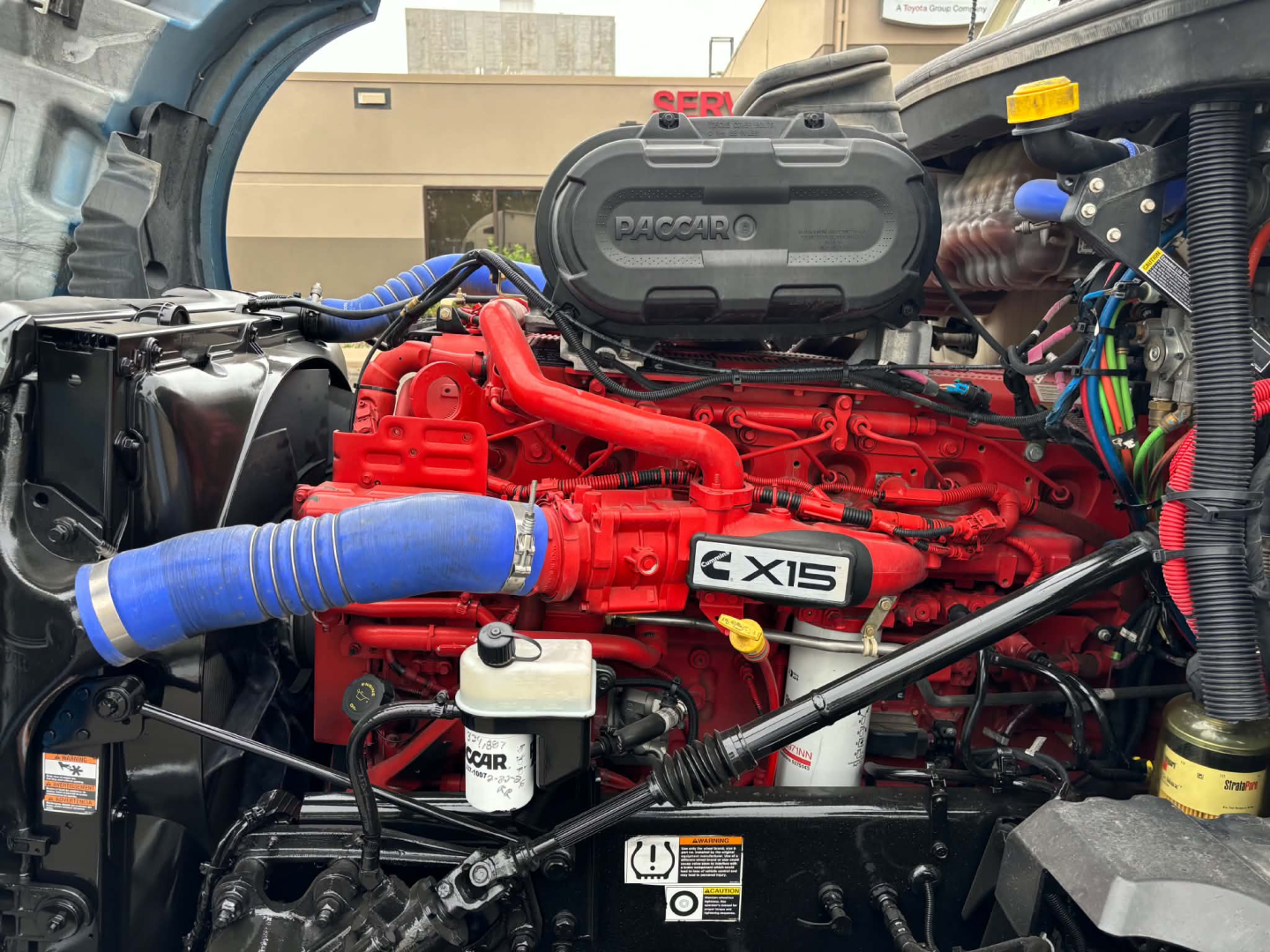 Kenworth T680 engine bay showing red Cummins X15 engine with blue intercooler hose, air intake, and various reservoirs and wiring.