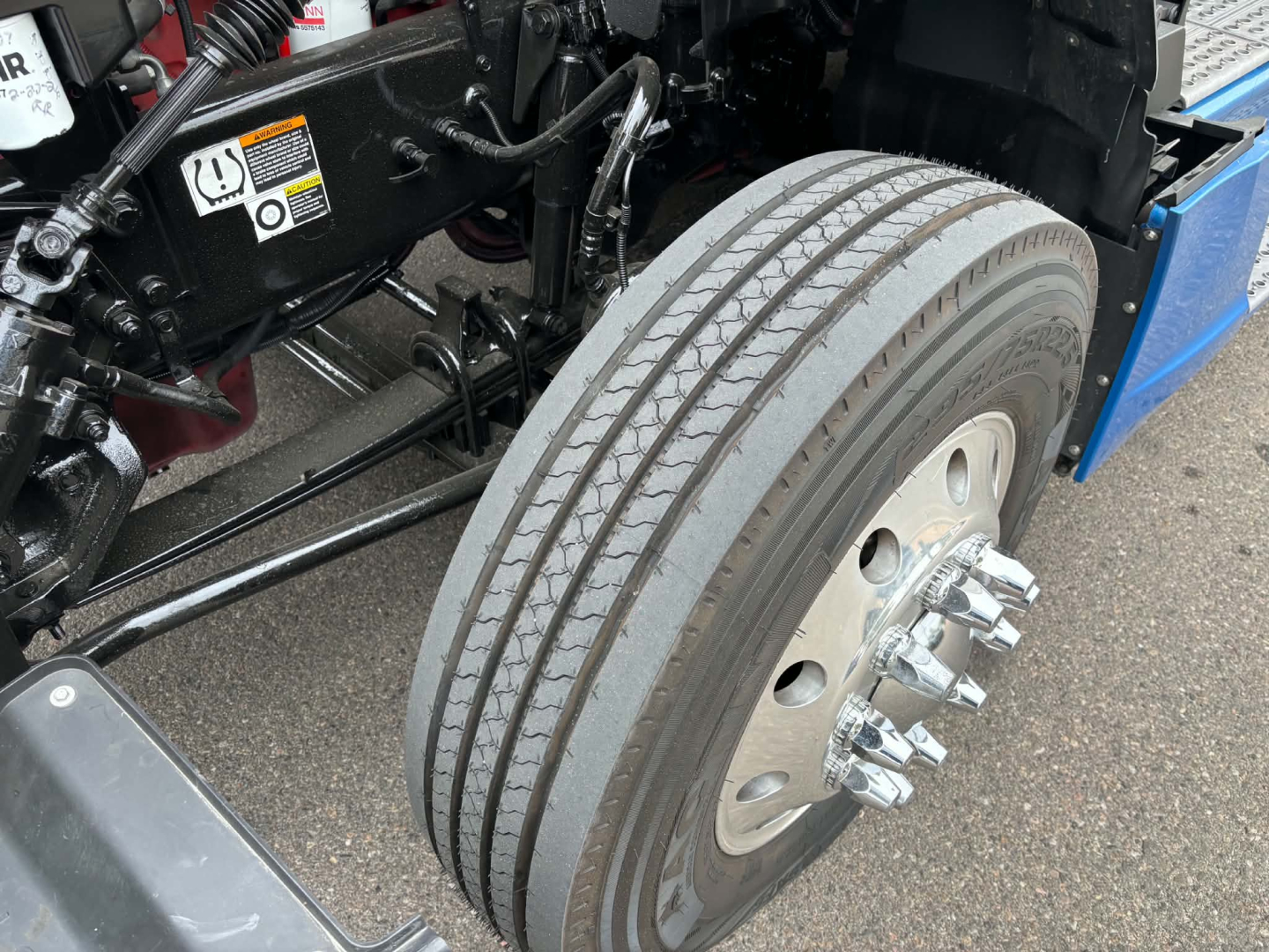 Kenworth T680 front wheel area showing dual rear tires, wheel studs, and part of the chassis with a blue fender; visible tires and rim bolts.