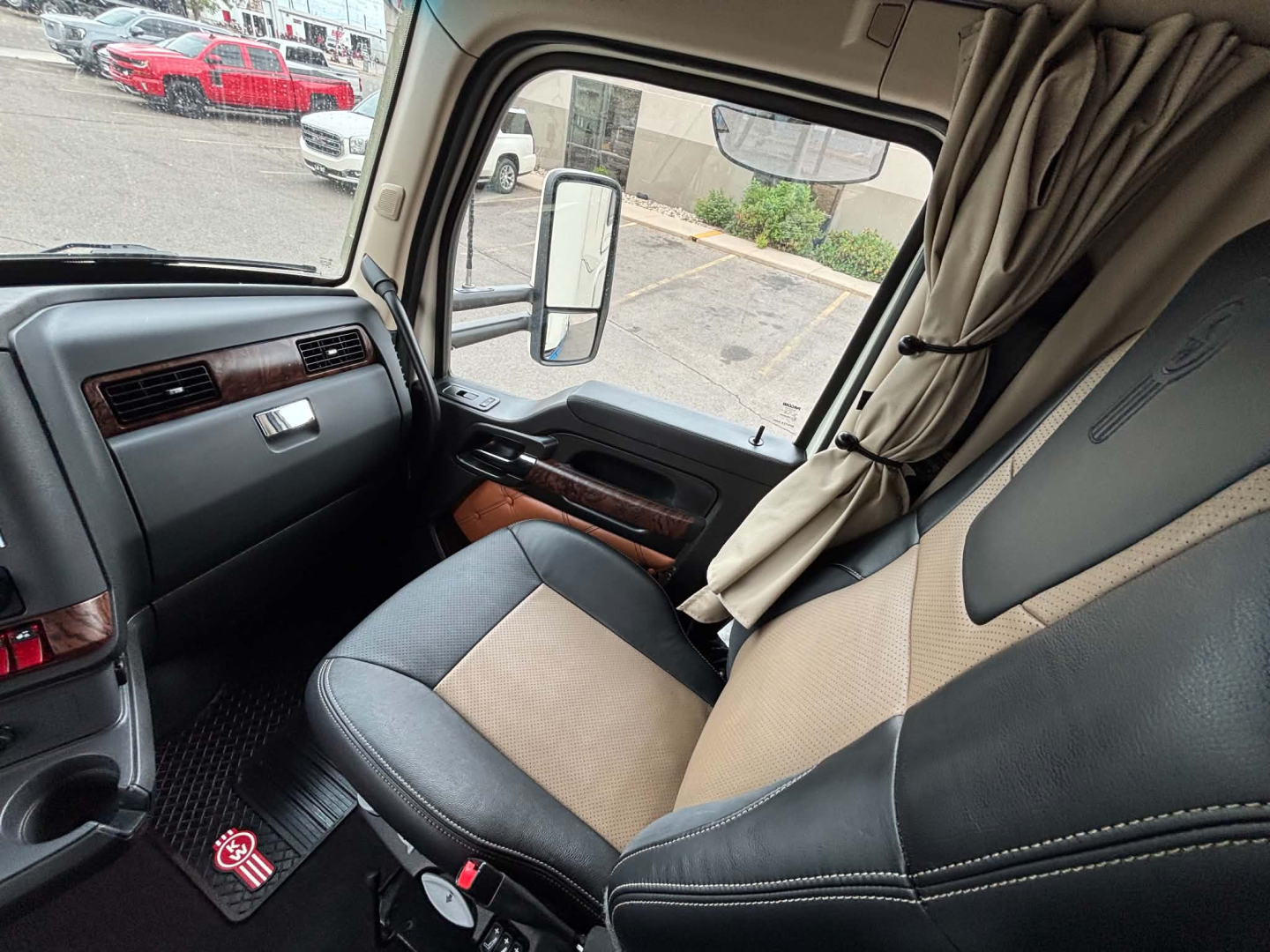 Kenworth T680 interior cab view showing driver seat and dash with window and side mirror; beige and black seat upholstery; curtain partition; dashboard trim.