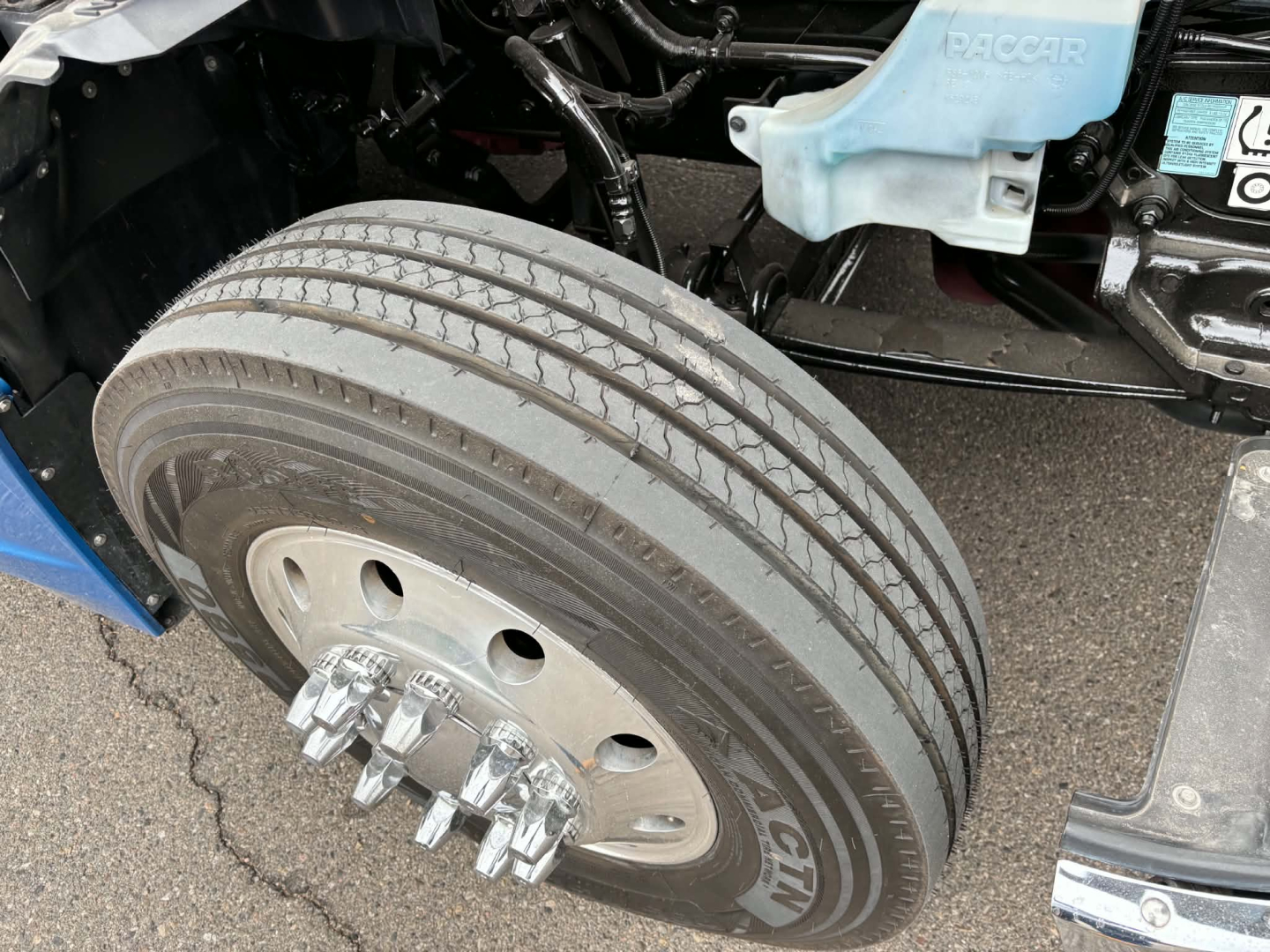 Kenworth T680 wheel and front suspension area showing dual rear axles with large steer tire and lug nuts, undercarriage details, blue DEF reservoir cap visible.