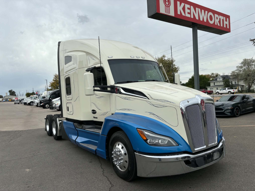 Kenworth T680 truck in showroom lot. Front grille with Kenworth badge, two-tone white and blue cab, side mirrors, and sign reading KENWORTH in background.