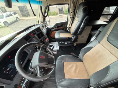 Truck cab interior of a Kenworth T680: steering wheel, dashboard, driver seat, passenger seat, center console, and door panel; visible upholstery in tan and black, multiple control panels, and gear lever.
