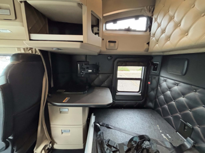 Truck cab interior of a 2024 Kenworth T680 showing sleeper area with quilted headboard, overhead storage, side window, and a fold-out desk/table.