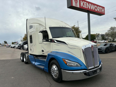 Kenworth T680 truck in showroom lot. Front grille with Kenworth badge, two-tone white and blue cab, side mirrors, and sign reading KENWORTH in background.
