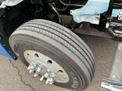 Kenworth T680 wheel and front suspension area showing dual rear axles with large steer tire and lug nuts, undercarriage details, blue DEF reservoir cap visible.