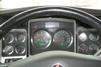 Kenworth truck dashboard view from steering wheel showing analog gauges, center digital display, and various indicator lights.