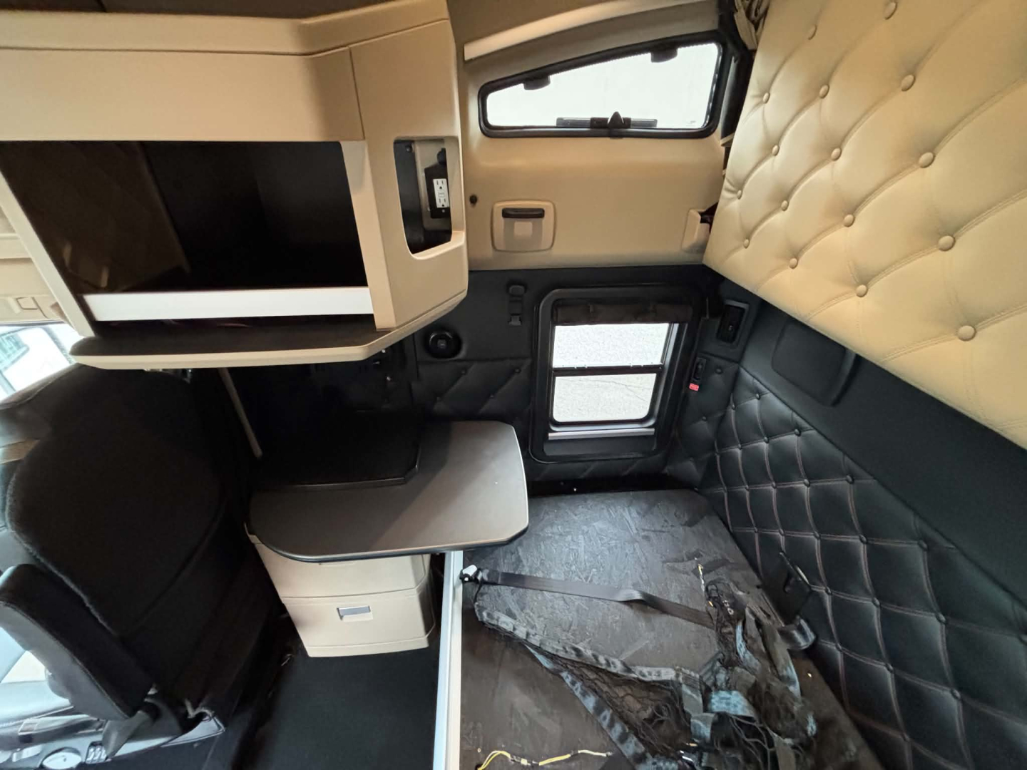Interior cab of a 2024 Kenworth T680 sleeper showing quilted black side panels, overhead cabinets, a small desk area, and a windowed door.