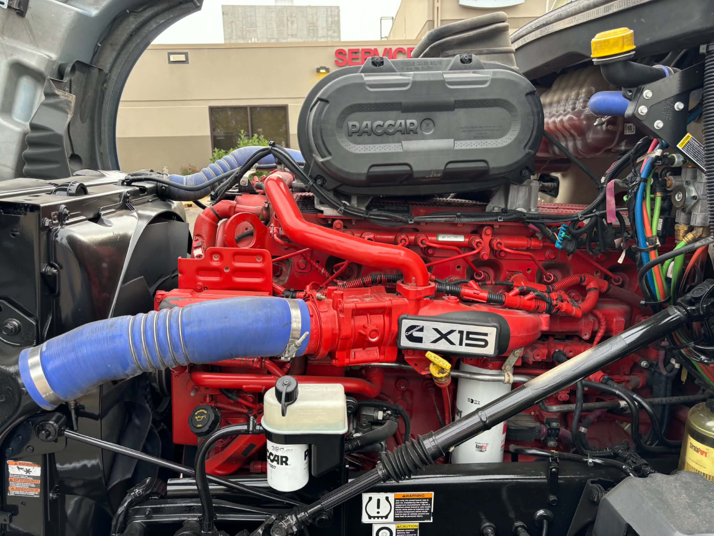 Kenworth T680 engine bay showing Cummins X15 red engine and blue intercooler hose, with black air intake housing and various wiring harnesses.