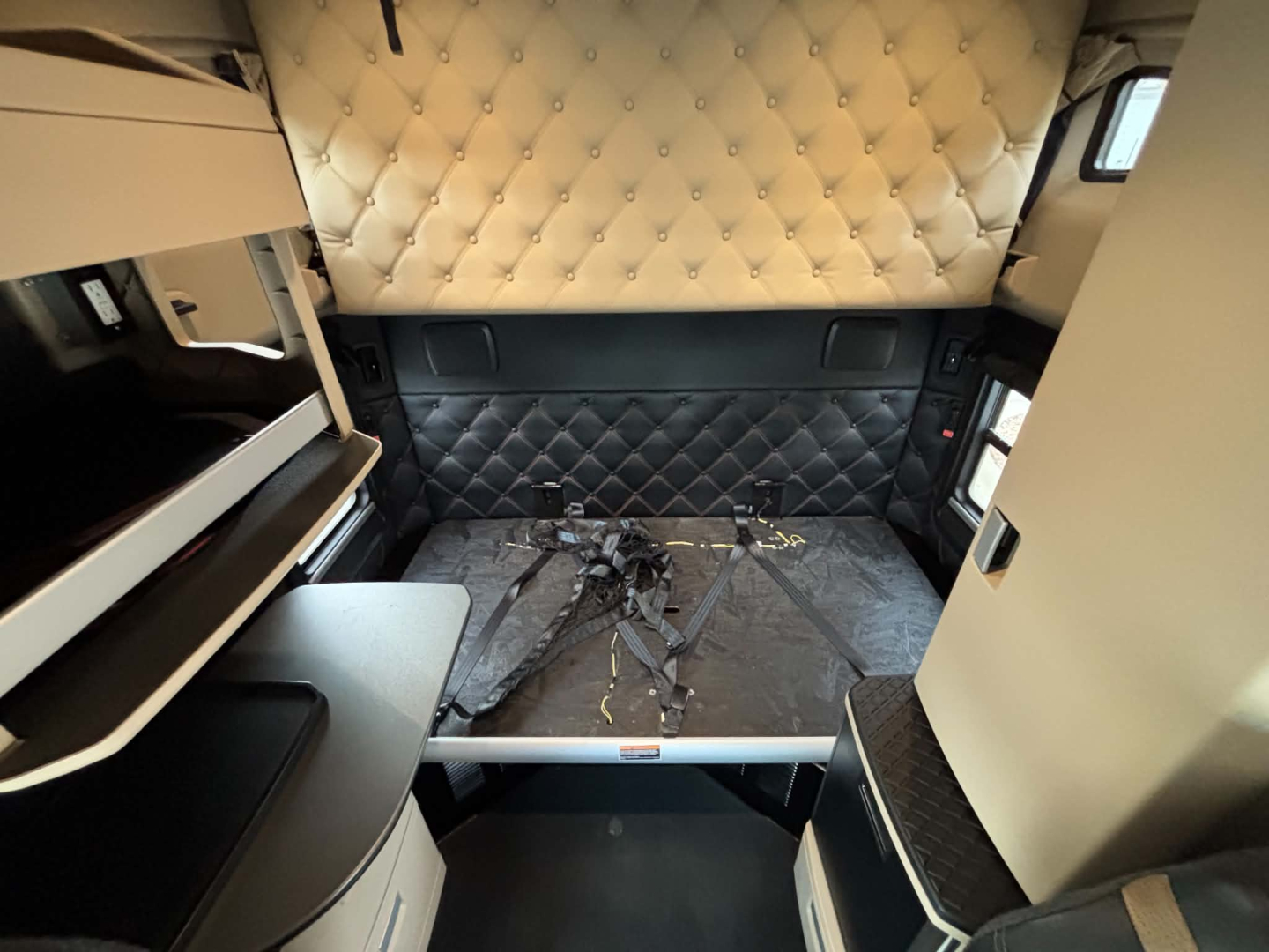 Interior of a 2024 Kenworth T680 sleeper cab showing quilted diamond headliner and side panels, with mounted seat area and cargo area hatch near the cab.