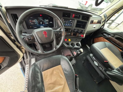 Interior view of a Kenworth T680 cab showing steering wheel with red emblem, dashboard, and driver's seat; center console with controls; brown and black leather seats.