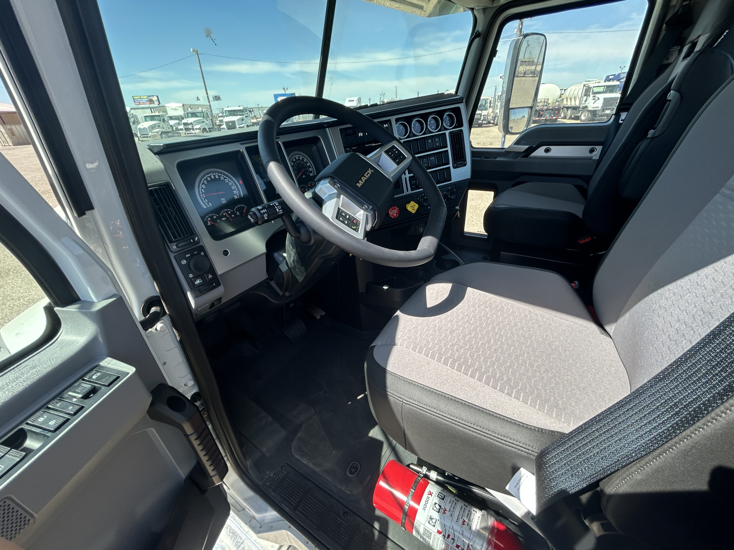 Cab interior of a 2027 Mack GR64FT view of the steering wheel and driver seat with dashboard gauges and door controls visible.