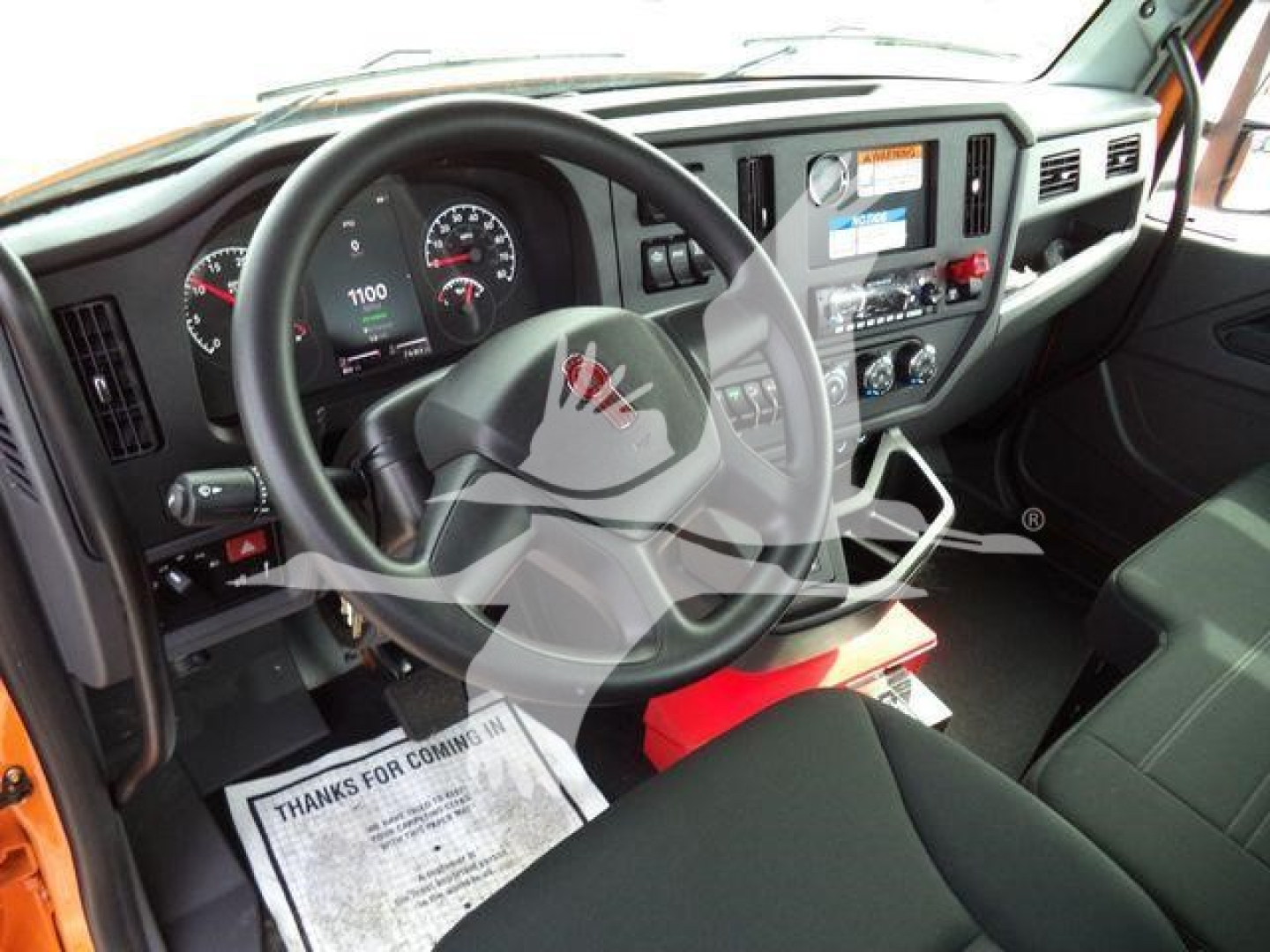 Truck cab interior view of a 2025 Kenworth T280 with steering wheel, dashboard gauges, center console, and orange exterior trim visible from the driver's seat.