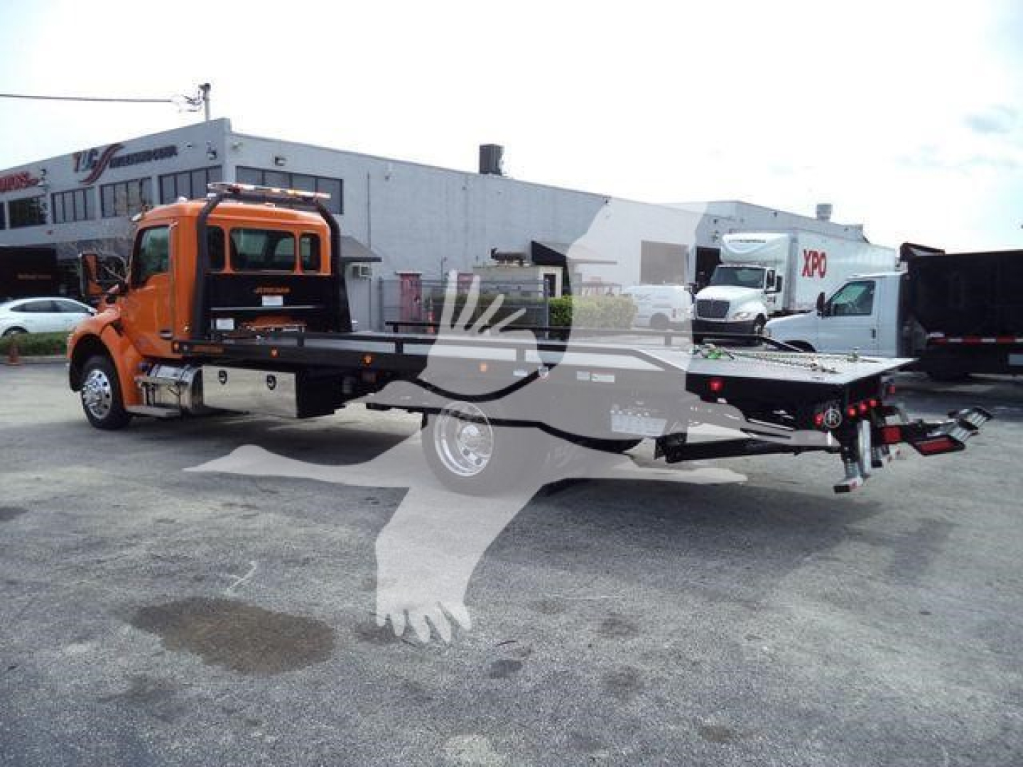 Flatbed rollback tow truck in orange with long flatbed and hydraulic winch, dealership lot in background.