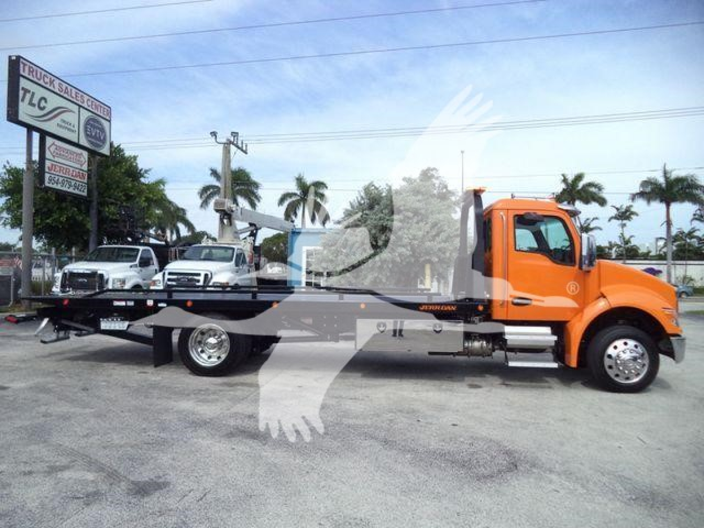 Kenworth T280 rollback tow truck in orange cab with flatbed, dealership lot in background, palm trees, and light blue sky.