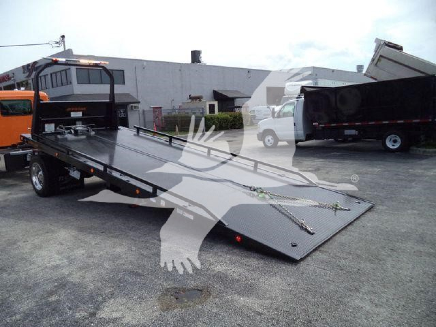 Kenworth T280 rollback truck with open flatbed ramp in foreground; orange and white trucks in background; visible text on image not legible.