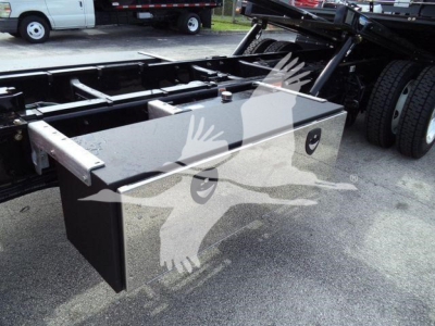 Flatbed area of a 2025 Kenworth T280 truck showing a large rectangular aluminum toolbox mounted on the frame near the rear, with chassis rails and wheel in the background.
