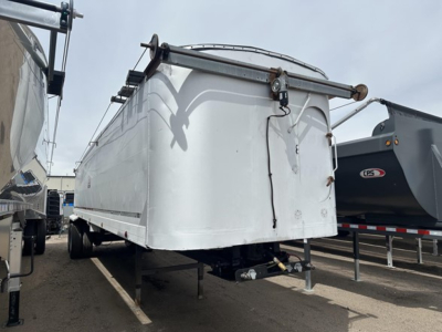 Frameless aluminum end dump trailer showing front nose and hitch area with rounded corners, white surface, and hydraulic lines visible beneath.