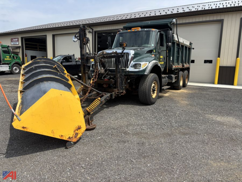 International WorkStar 7600 dump truck with snow plow attachment in front, dark green cab, large yellow plow blade, dealership backdrop.