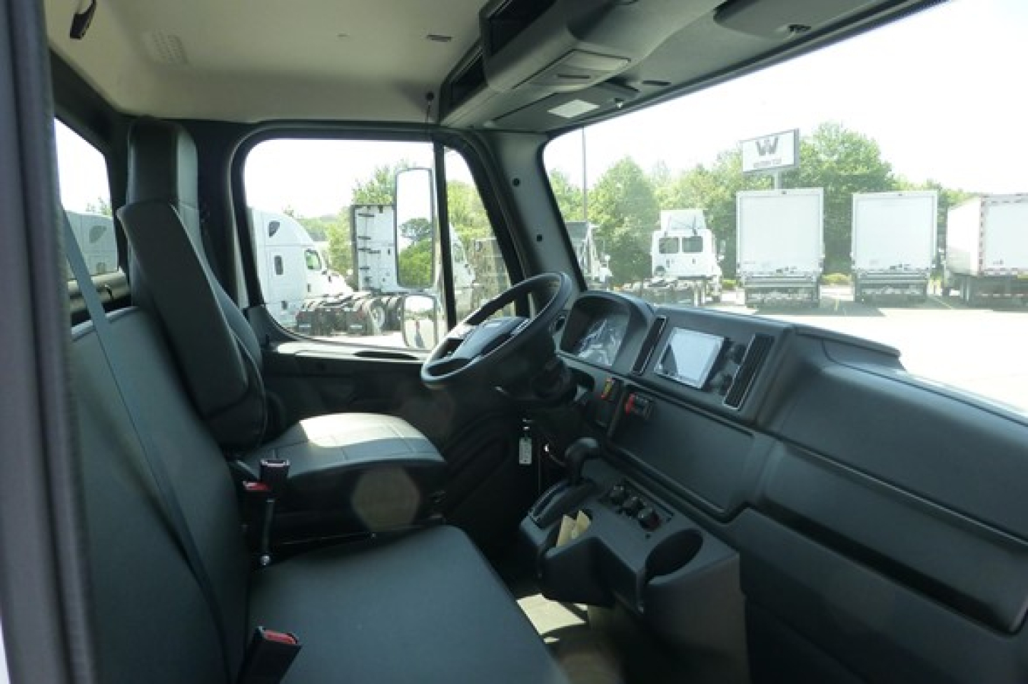 Cab and chassis truck cockpit interior showing steering wheel, dashboard with digital display, and seats; window view reveals trucks outside.