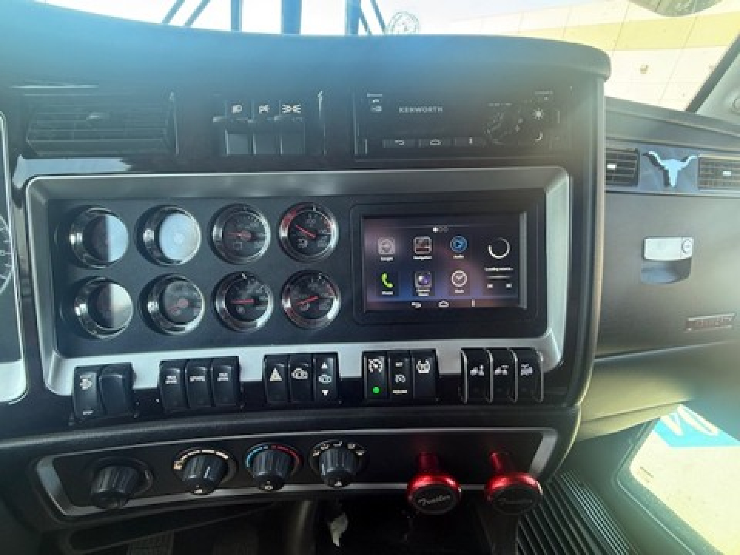 Truck dashboard and center console with multiple gauges, a touchscreen infotainment display, and control switches; Kenworth branding on the dash.