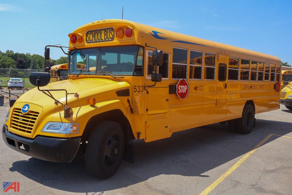 Yellow school bus from side/front angle with 'SCHOOL BUS' sign on roof and number 5333, stop sign extended, blue sky.
