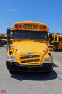 Yellow school bus front view showing front grille and headlights, with a blue bird emblem and 'SCHOOL BUS' sign on the roof and other buses in the background.