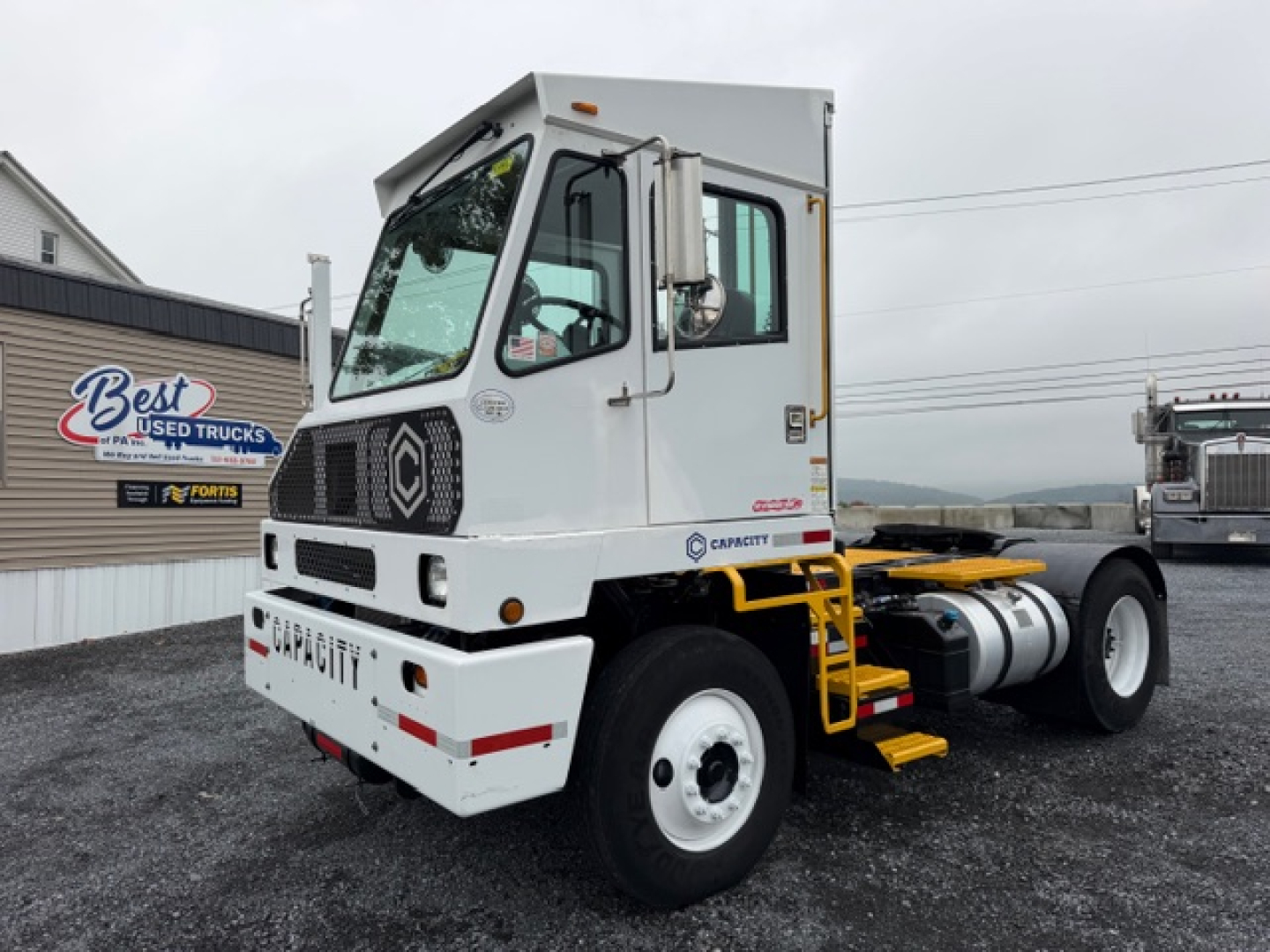 Capacity TJ5000 truck tractor in yard showing front and side, cab with white paint, black grille, and yellow steps; DOT capacity label on door area.