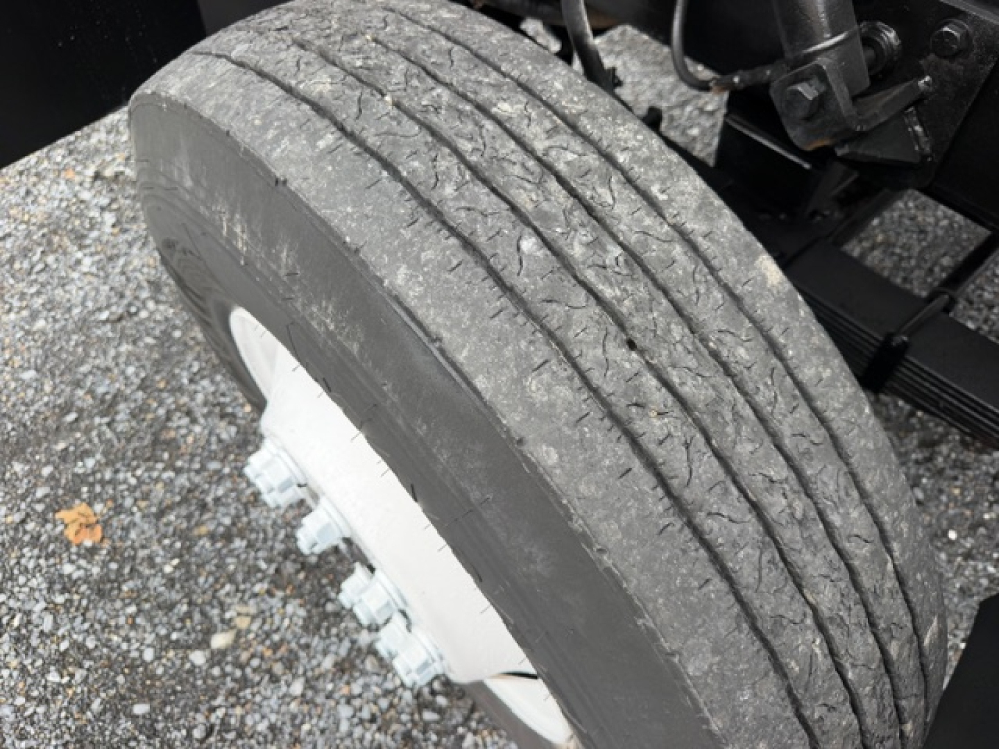 Wheel and tire of heavy-duty truck chassis with mud and gravel; visible bolts on hub; part of the axle and frame are in view.