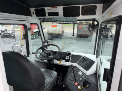 Truck cabin interior showing steering wheel, dashboard, and control levers with a windshield view to yard equipment.