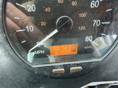 Dashboard gauge cluster showing mph and km/h scale with orange hour counter display; two control buttons labeled MODE and SET below the gauge; reflected hand in glass.