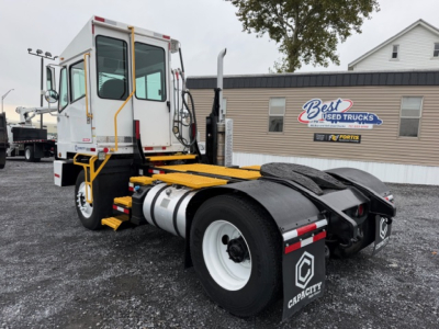 Truck chassis and cab with a large yellow platform and rear wheel area, fuel tank, and white cab; Best Used Trucks sign on building in background.