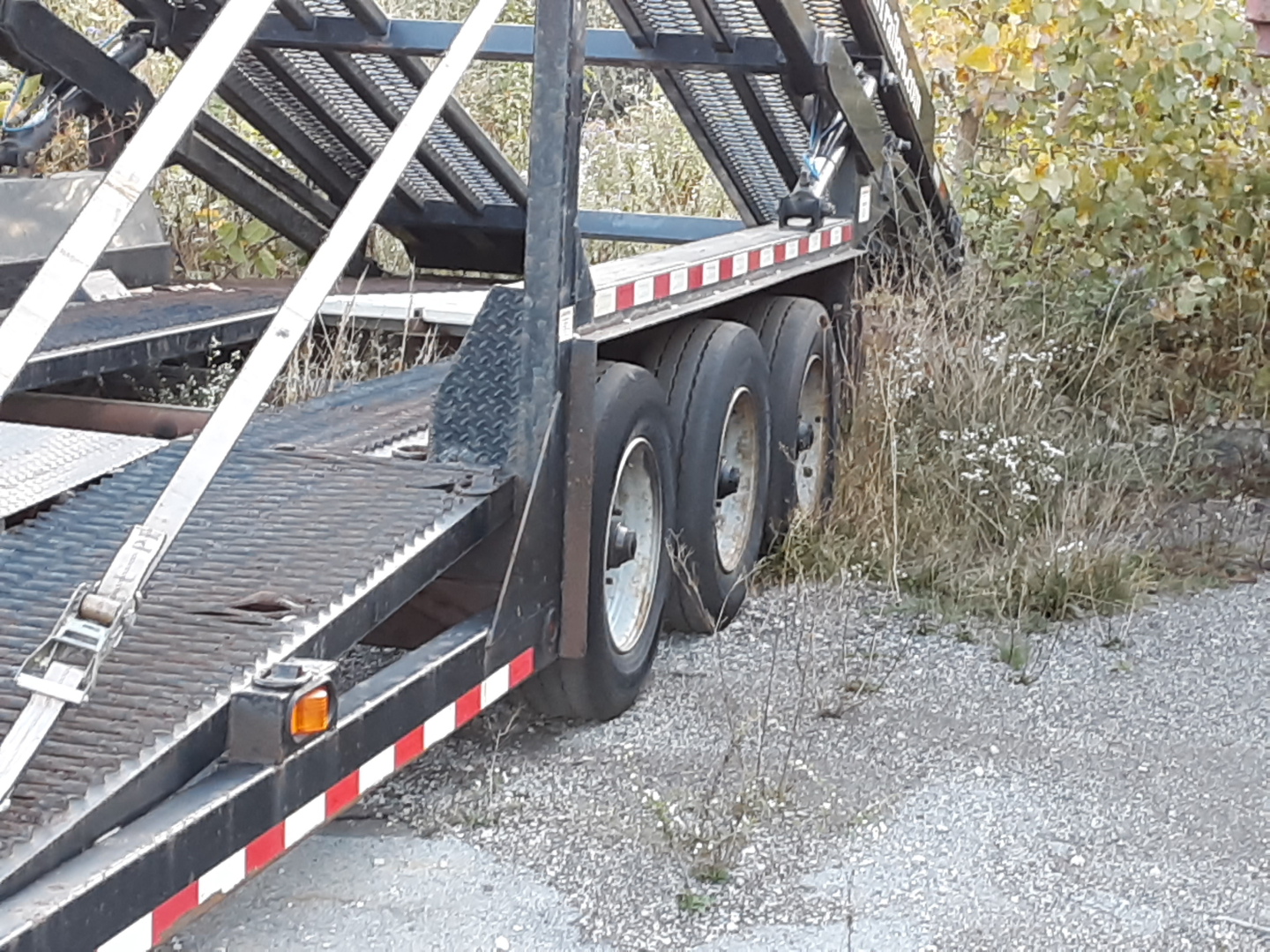 Trailer deck of a car carrier with multiple axles and black metal framework. Flatbed with diamond plate; white and red reflective safety tape along the sides. Some vegetation in the background.
