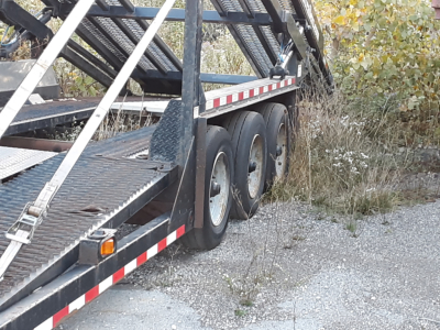 Trailer deck of a car carrier with multiple axles and black metal framework. Flatbed with diamond plate; white and red reflective safety tape along the sides. Some vegetation in the background.