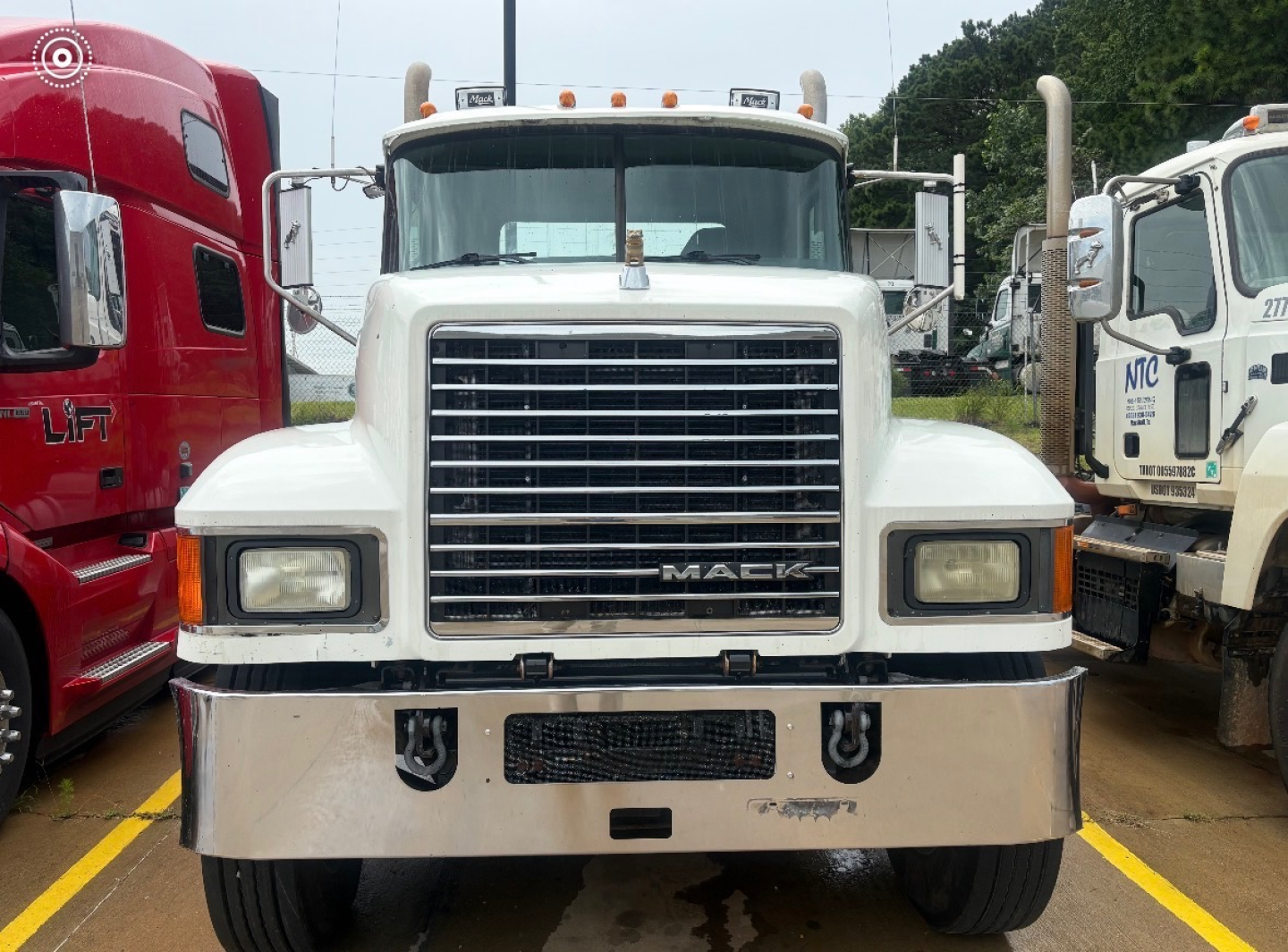 Mack CHU613 truck front view showing large chrome grille, rectangular headlights, and chrome bumper; white cab with side mirrors; other trucks visible on either side.
