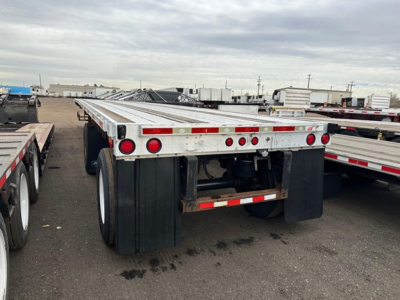 Flatbed trailer rear view showing dual axle, mud flaps, and aluminum deck with red taillights and reflectors.
