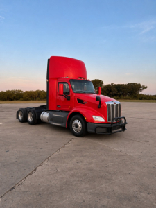 Peterbilt 579 conventional daycab tractor in red color, side view with sleeper cab raised, front grille, daytime running lights, chassis, and triple rear axles.
