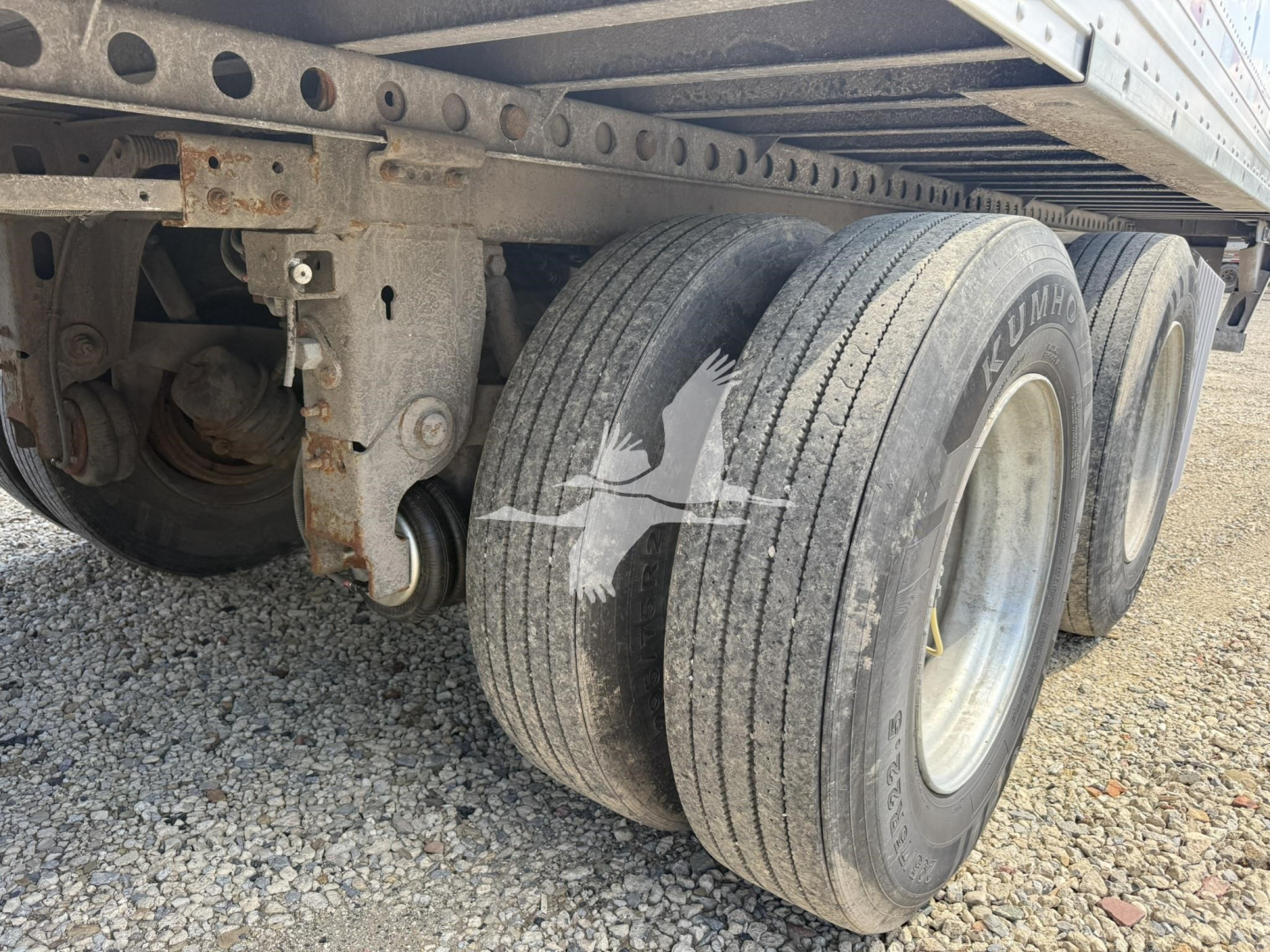Triple-axle trailer undercarriage showing dual rear tires and leaf spring suspension with rust-free frame. Visible: wheels, tires, axle housing, and frames.